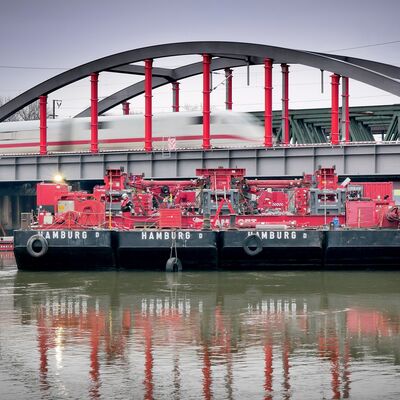 Schwimmende Arbeitsplattform mit Hubvorrichtung vor einer Eisenbahnüberführung.