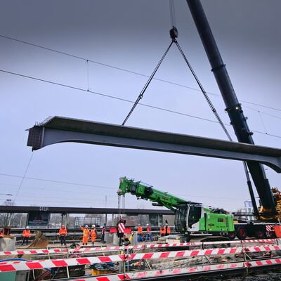 Großer Stahlträger an einem Mobilkran. Im Vordergrund Gleise und Bauarbeiter.