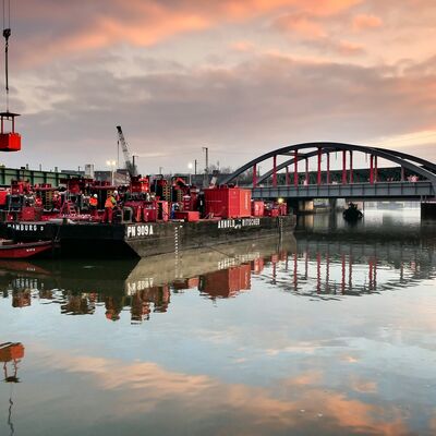 Links vorne eine schwimmende Arbeitsplattform vor einer Eisenbahnbrücke.