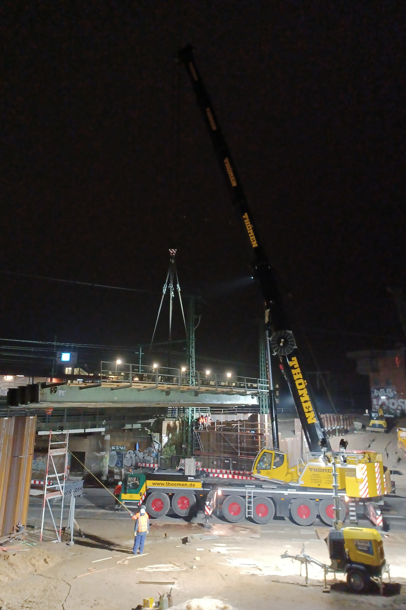 Mobilkran lädt nachts eine Hilfsbrücke von Schwerlasttransporter und hebt sie ein.