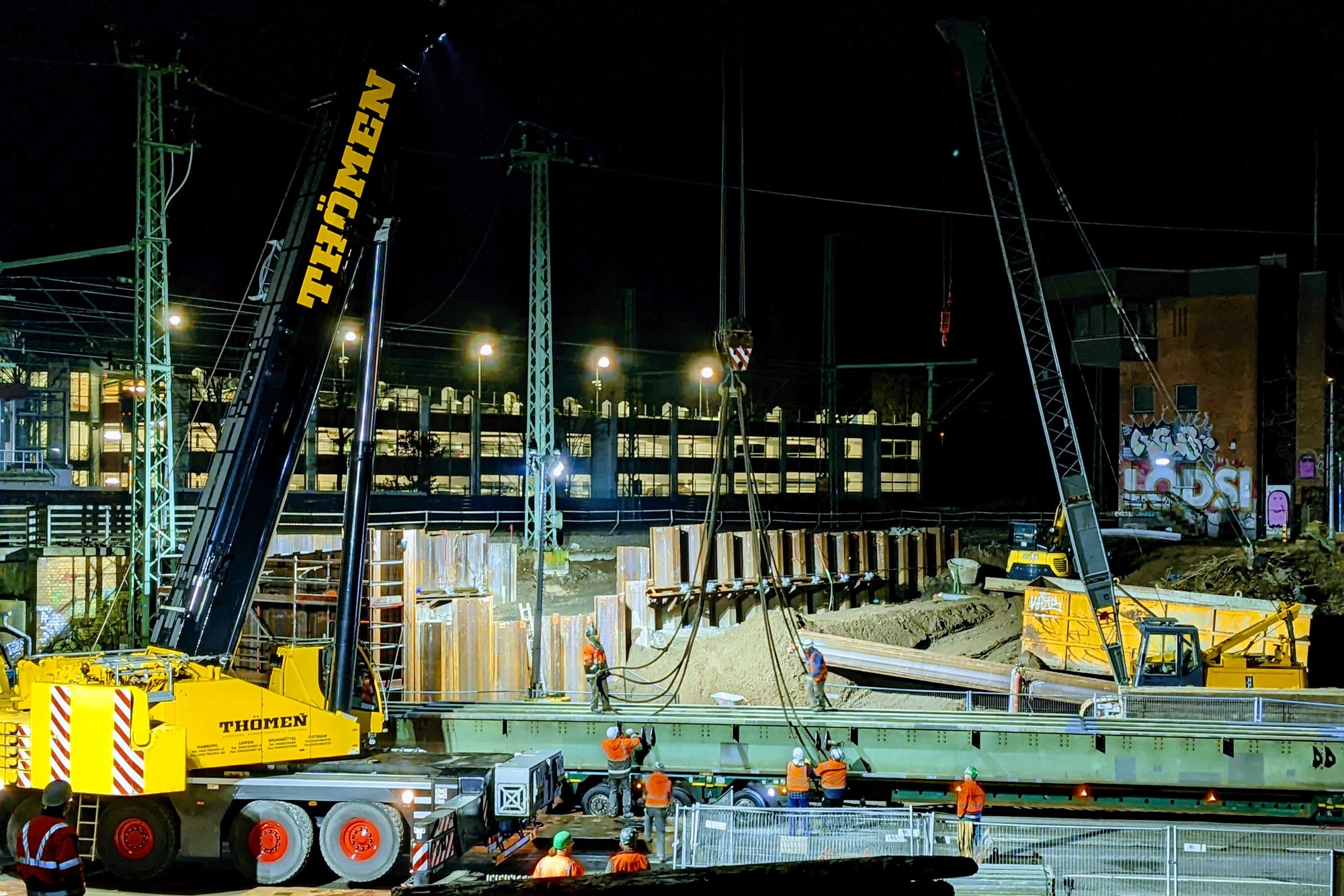 Ein Mobilkran hebt nachts eine Hilfsbrücke von einem Schwerlasttransporter.