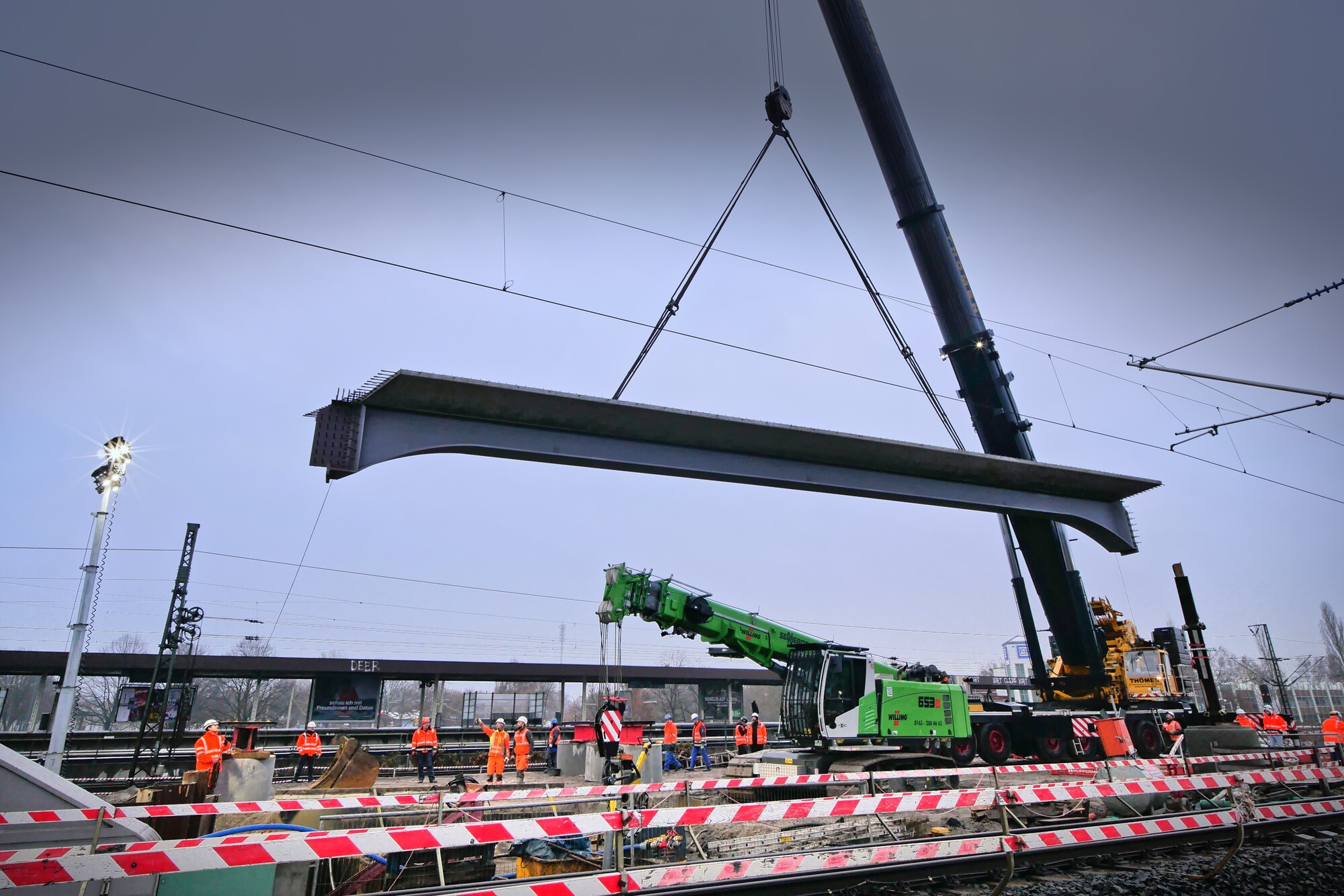 Großer Stahlträger an einem Mobilkran. Im Vordergrund Gleise und Bauarbeiter.